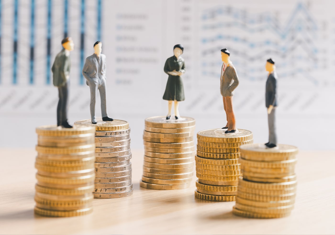 Miniature people on stacks of coins in front of financial charts.
